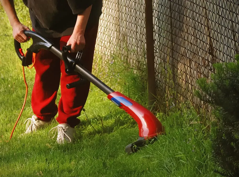 Worker in red pants using a string trimmer along a chain-link fence
