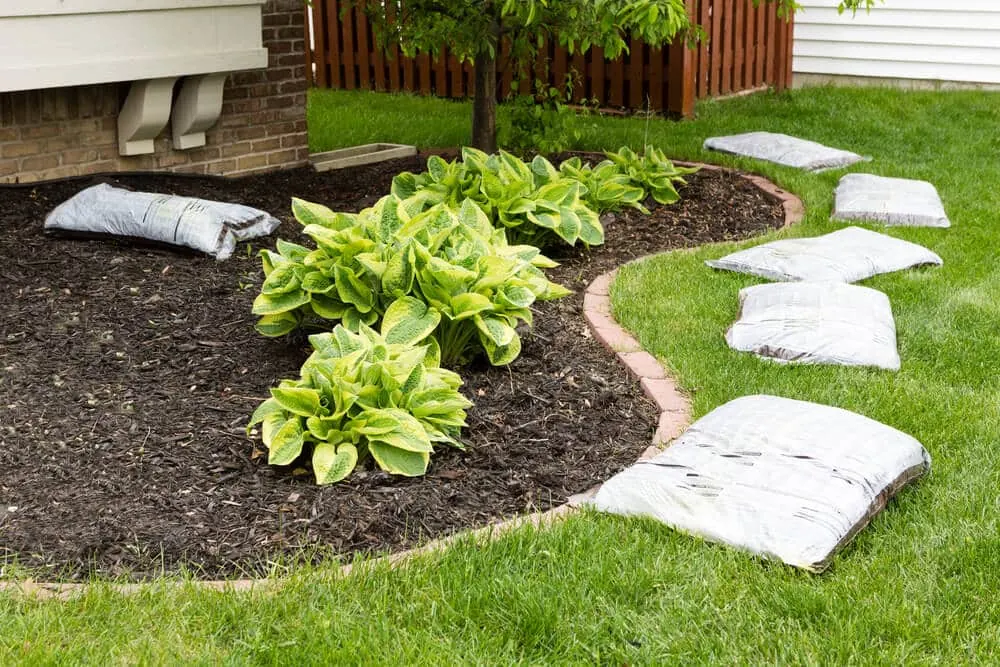 Freshly mulched curved bed with hostas, edged by bricks, with unopened mulch bags lined up on the lawn