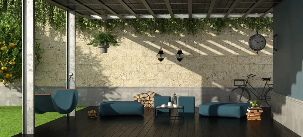 Covered lounge with blue outdoor seating, hanging lanterns, and vines trailing from a slatted roof beside a stone wall