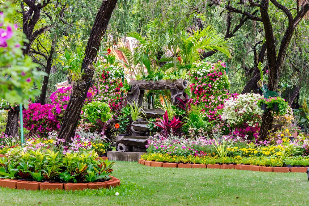 Flower garden with pink and red blooms, brick-edged beds, and a small lawn under trees